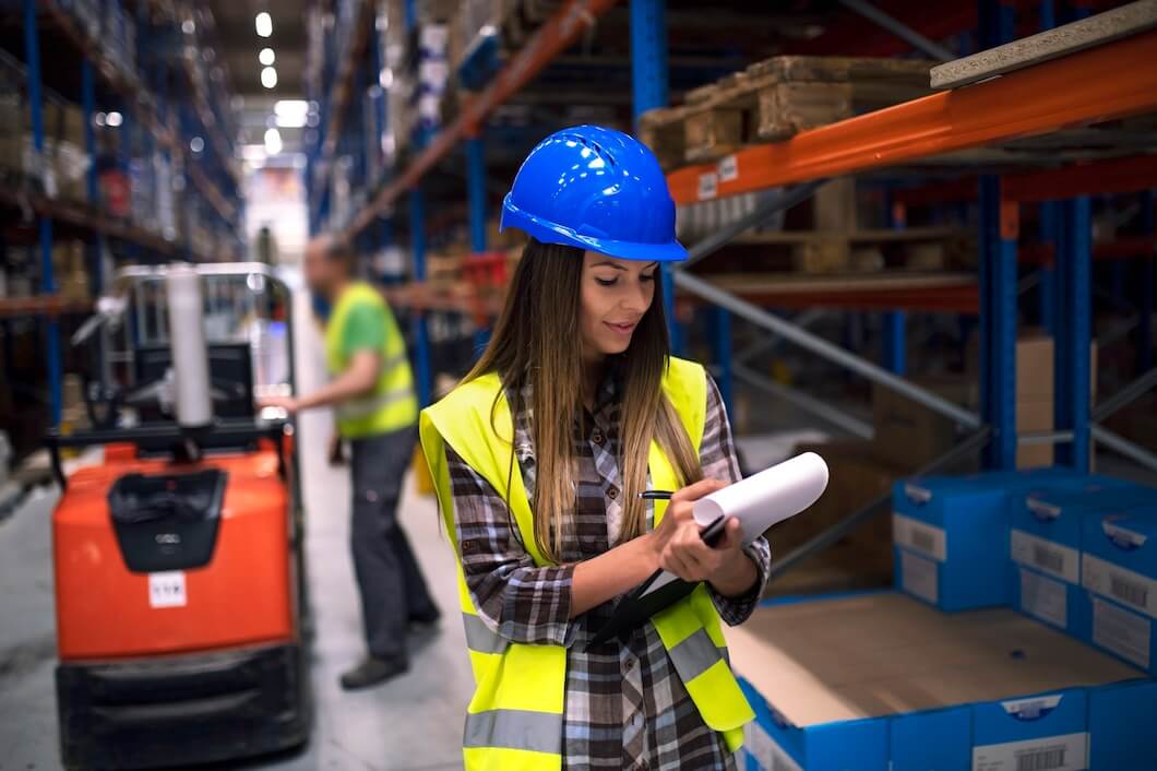  portrait-female-warehouse-worker-checking-inventory-storage-department-while-her-coworker-operating-forklift